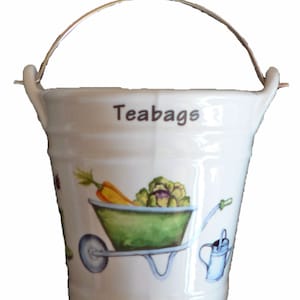 May include: White ceramic teabag holder shaped like a bucket with a metal handle. The bucket has a green wheelbarrow with vegetables and a watering can printed on it. The text "Teabags" is printed on the front of the bucket.