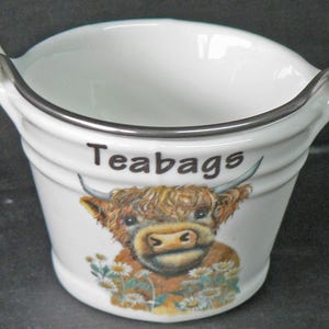 May include: White ceramic teabag holder with a Highland cow illustration and the word "Teabags" printed on the front. The bucket-shaped container has a silver rim and handles. Daisies are at the bottom of the cow illustration.