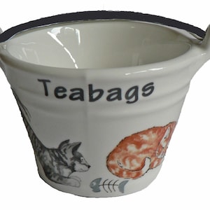 May include: White ceramic tea bag holder shaped like a bucket with a black handle. The bucket has a black and white cat and an orange and white cat on the side. The text "Teabags" is printed on the front of the bucket.