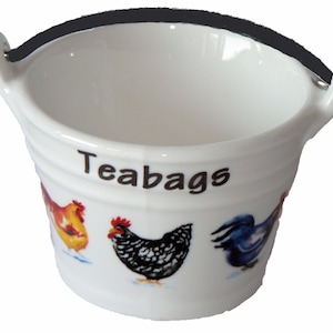 May include: White ceramic tea bag holder shaped like a bucket with a black handle. The bucket has a brown inscription that says "Teabags" and a colourful illustration of chickens.