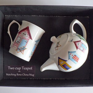 May include: A white teapot and mug set with a beach hut design. The teapot is two-cup and the mug is bone china. The text on the box reads "Two cup Teapot with Matching Bone China Mug".