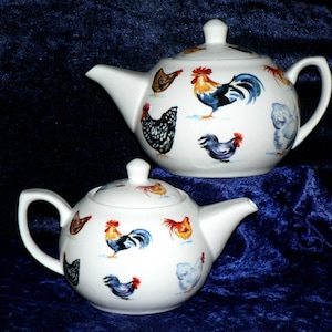 May include: Two white ceramic teapots with lids, decorated with colourful rooster and hen illustrations. The teapots have curved spouts and handles. The background is a dark blue velvet fabric.