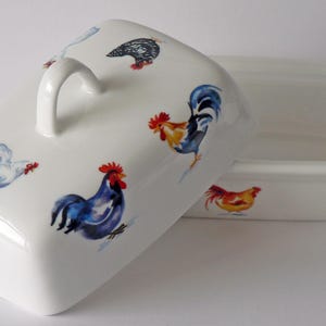 May include: White ceramic butter dish with a lid featuring colorful rooster and hen illustrations. The lid has a curved handle. The dish is rectangular with rounded edges. The rooster and hen designs are in shades of red, blue, yellow, and brown.