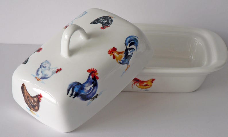 May include: White ceramic butter dish with a lid featuring colorful rooster and hen illustrations. The lid has a curved handle. The dish is rectangular with rounded edges. The rooster and hen designs are in shades of red, blue, yellow, and brown.