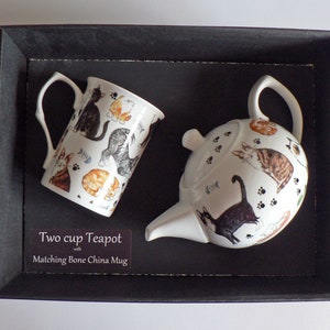 May include: A white two-cup teapot and matching bone china mug with a cat design. The teapot and mug feature a variety of cats in different poses, including a black cat, a tabby cat, and a ginger cat. The background of the teapot and mug is white with paw prints.