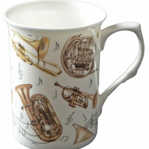 May include: A white ceramic mug featuring a design of musical instruments, including a trumpet, trombone, tuba, and French horn. The instruments are gold and brown and are surrounded by musical notes.