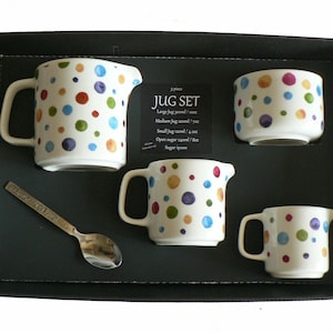 May include: A white ceramic jug set with a colourful polka dot pattern. The set includes a large jug, a medium jug, a small jug, an open sugar bowl, and a sugar spoon. The set is packaged in a black box with the text "JUG SET" printed on it.
