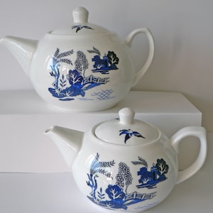 May include: Two white ceramic teapots with blue and white willow pattern designs. The larger teapot is in the background and the smaller teapot is in the foreground.