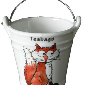 May include: White ceramic tea bag holder shaped like a bucket with a silver handle. The bucket has a cartoon fox with big eyes and a bushy tail printed on it. The word "Teabags" is printed on the front of the bucket.