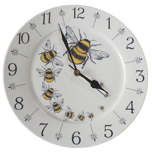 May include: A white ceramic clock with black numbers and hands. The clock face features a design of bees in a circle around the clock face. The bees are yellow and black with black wings.