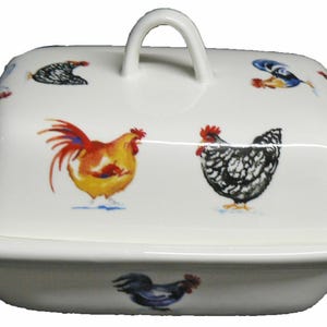 May include: White ceramic butter dish with a lid featuring colorful rooster illustrations. The dish has a rectangular base and a curved lid with a handle. The roosters are in shades of red, yellow, and black.