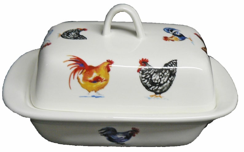 May include: White ceramic butter dish with a lid featuring colorful rooster illustrations. The dish has a rectangular base and a curved lid with a handle. The roosters are in shades of red, yellow, and black.