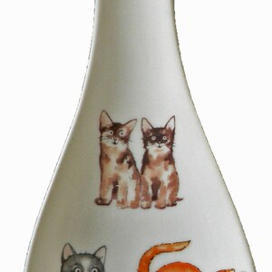 May include: White ceramic spoon rest with a decorative design of four different cats. The cats are in various poses and colours, including grey, orange, and brown. The spoon rest is approximately 20 cm long.