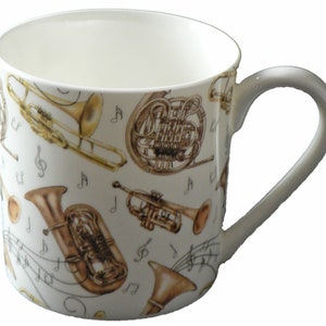 May include: A white ceramic mug featuring a design of musical instruments, including a trumpet, trombone, French horn, and tuba. The instruments are illustrated in gold and brown hues against a white backdrop.