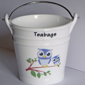 May include: White ceramic tea bag holder shaped like a bucket with a silver handle. The bucket has a blue and purple owl design with the word "Teabags" printed on the side.