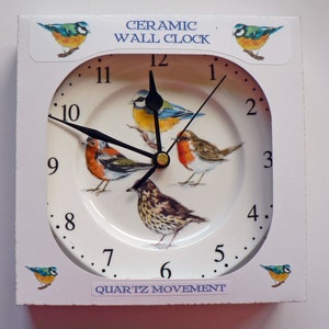 Pu&ograve; includere: Un orologio da parete in ceramica bianca con una faccia quadrata e una faccia rotonda. La faccia dell'orologio presenta un design di uccelli e un movimento al quarzo. L'orologio &egrave; in una scatola bianca con le parole "Orologio da parete in ceramica" e "Movimento al quarzo" stampate su di essa.