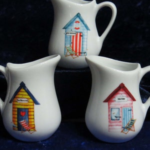 May include: Three white ceramic milk jugs with colourful beach hut designs. Each jug features a different beach hut with a unique colour scheme and details. The huts have signs that read "The Beach Hut", "Sea View", and "Old Skool".