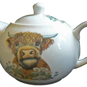May include: A white ceramic teapot with a lid and handle. The teapot is decorated with a Highland cow illustration, featuring brown fur, horns, and a daisy field. A delightful kitchen accessory.