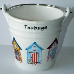 May include: White ceramic teabag holder shaped like a bucket with a silver handle. The bucket has a colourful illustration of three beach huts with red, yellow, blue and white stripes. The text "Teabags" is printed on the front of the bucket.