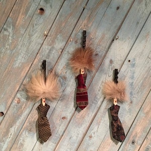 May include: Three handmade ornaments featuring a brown, black, and red tie with a fuzzy brown hairpiece on top. The ornaments are hanging from a wooden surface.