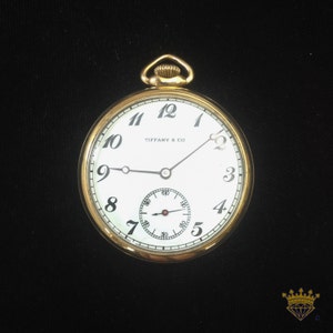 K&ouml;nnte beinhalten: Eine goldene Taschenuhr mit wei&szlig;em Zifferblatt und schwarzen r&ouml;mischen Ziffern. Die Uhr hat einen schwarzen Sekundenzeiger und den Text "Tiffany & Co." auf dem Zifferblatt.