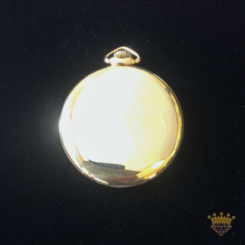 K&ouml;nnte beinhalten: Eine goldene Taschenuhr mit einem runden Geh&auml;use und einem schlichten Design. Die Uhr hat eine glatte, polierte Oberfl&auml;che und einen kleinen Knopf oben.