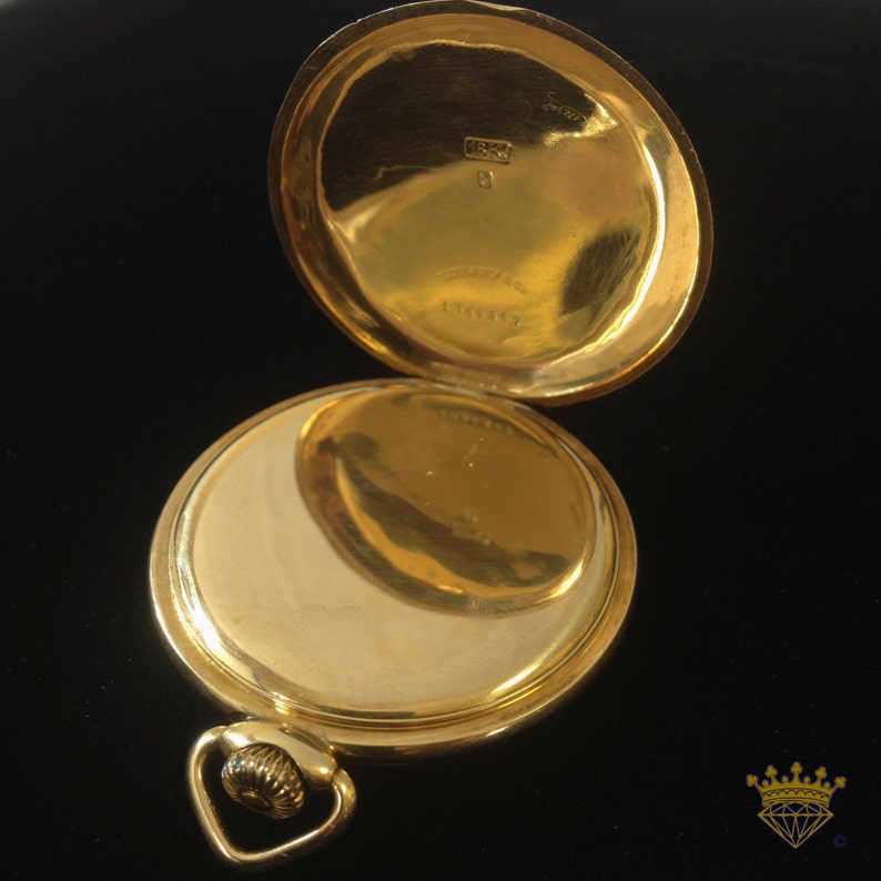 K&ouml;nnte beinhalten: Eine goldene Taschenuhr mit einem aufklappbaren Deckel, der das Innenleben der Uhr freilegt. Die Uhr hat ein dekoratives Design auf der R&uuml;ckseite und eine kleine Schlaufe zum Anbringen einer Kette.