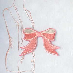 Op de afbeelding: Een roze geborduurde strikpatch met een lichtgrijze kern, tegen een witte achtergrond met een schets van een paspop. De strik heeft een gedetailleerd steekpatroon en puntige uiteinden, perfect voor het verfraaien van kleding of accessoires.