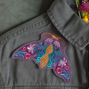 Peut inclure: Un écusson brodé en forme de papillon de nuit aux couleurs vives, avec des détails violets, turquoise et dorés, est fixé sur une veste grise. L'écusson présente des motifs floraux et géométriques. Un petit bouquet de fleurs est glissé dans le col de la veste.