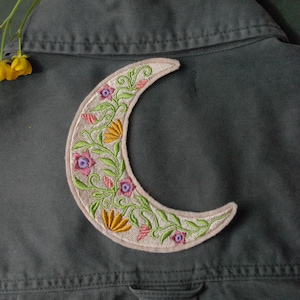 May include: Embroidered crescent moon patch with floral design in pink, yellow, and green. The patch is on a dark green fabric, with a bouquet of colourful flowers in the background. The patch is a decorative element.