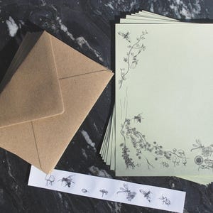 May include: A stack of pale green stationery sheets featuring black line art of bees, flowers, and insects along the left and bottom edges, accompanied by a brown kraft paper envelope and a strip of matching insect-themed stickers.