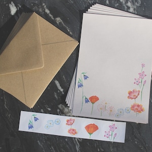 Peut inclure: Une pile de papier à lettres de couleur claire ornée d'une bordure florale à l'aquarelle avec des fleurs bleues, orange et roses, accompagnée d'un ensemble d'enveloppes en papier kraft brun et d'une bande horizontale d'autocollants assortis.