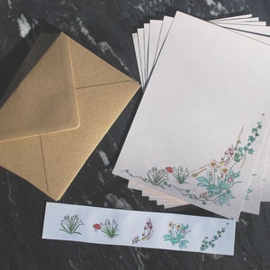 Peut inclure: Une pile de feuilles de papier à lettres blanc cassé ornées d'une illustration botanique de vignes vertes et de petites fleurs dans le coin inférieur droit, avec une enveloppe marron assortie et une bande horizontale aux motifs floraux.