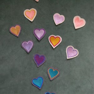 May include: A collection of twelve small heart-shaped embroidered patches in various colours including pink, purple, gold, and blue. Each heart features a stitched border and a textured thread finish, arranged in a scattered pattern across the frame.