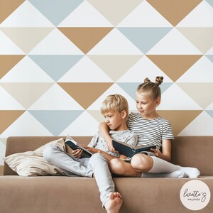 Può includere: Due bambini leggono un libro insieme su un divano marrone. Lo sfondo presenta un design geometrico della carta da parati con triangoli bianchi, beige, azzurri e marroni. Un cuscino poggia sul divano. Il marchio "Luvette's Wallpaper" è nell'angolo in basso a destra.