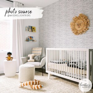 May include: A nursery room featuring white walls with black horizontal dashed line wallpaper, a white wooden crib, a light gray armchair, a textured white ottoman, a woven lion wall hanging, and a white cylindrical side table with a doll.