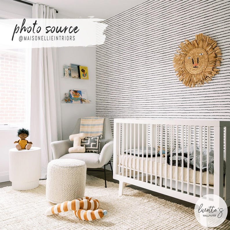 May include: A nursery room featuring white walls with black horizontal dashed line wallpaper, a white wooden crib, a light gray armchair, a textured white ottoman, a woven lion wall hanging, and a white cylindrical side table with a doll.