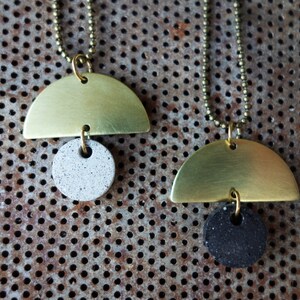 May include: Two gold half-circle pendants with a round concrete charm. The charms are different colors, one is white and the other is black.