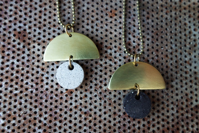 May include: Two gold half-circle pendants with a round concrete charm. The charms are different colors, one is white and the other is black.
