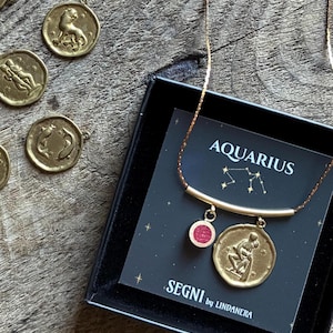 Può includere: Collana con pendente Acquario color oro con un ciondolo rotondo e un piccolo ciondolo rosso. La collana è esposta in una scatola nera con la scritta "AQUARIUS" e "SEGNI by LINDANERA" stampata sulla carta. Sono visibili altri pendenti dello zodiaco.