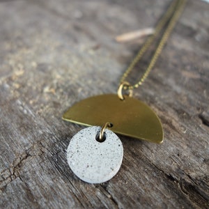 May include: A gold and gray necklace with a half-circle pendant and a round pendant. The necklace is on a wooden surface.