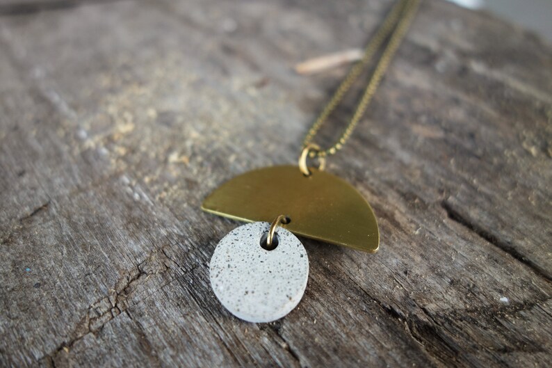 May include: A gold and gray necklace with a half-circle pendant and a round pendant. The necklace is on a wooden surface.