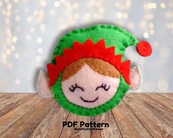 Könnte beinhalten: Ein Weihnachts-Elfen-Anhänger aus Filz mit grünem Hut, rotem Besatz und einem lächelnden Gesicht. Der Anhänger befindet sich auf einer Holzoberfläche. PDF Pattern thelittlefeltflower