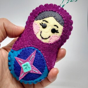 May include: A purple felt doll with a pink and blue star on its skirt. The doll has a smiling face with black eyes and a black nose.
