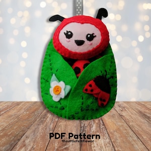 Puede incluir: Un adorno de mariquita de fieltro con un diseño de hoja verde. La mariquita tiene un cuerpo rojo con manchas negras y una cara blanca con ojos negros. El adorno es perfecto para una guardería o una habitación infantil.