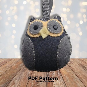 May include: A gray felt owl ornament with yellow felt accents around the eyes and beak. The owl has black button eyes and is hanging from a gray ribbon with white circles. The owl is sitting on a wooden surface. PDF Pattern thelittlefeltflower