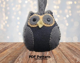 May include: A gray felt owl ornament with yellow felt accents around the eyes and beak. The owl has black button eyes and is hanging from a gray ribbon with white circles. The owl is sitting on a wooden surface. PDF Pattern thelittlefeltflower