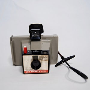 May include: A gray Polaroid Big Swinger 2000 camera with a black lens and a red button. The camera has a black strap attached to it.