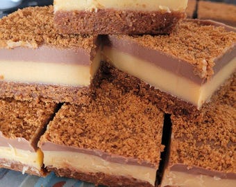 May include: A close-up of a stack of homemade layered dessert bars. The bars have a crumbly cookie crust, a layer of caramel, and a layer of chocolate. The bars are arranged in a pyramid shape on a blue and white striped surface.