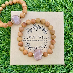 May include: Two wooden bead bracelets with a pink sunstone charm. The bracelets are in a brown box with the text "Cory Willa Handmade Aromatherapy Jewelry".
